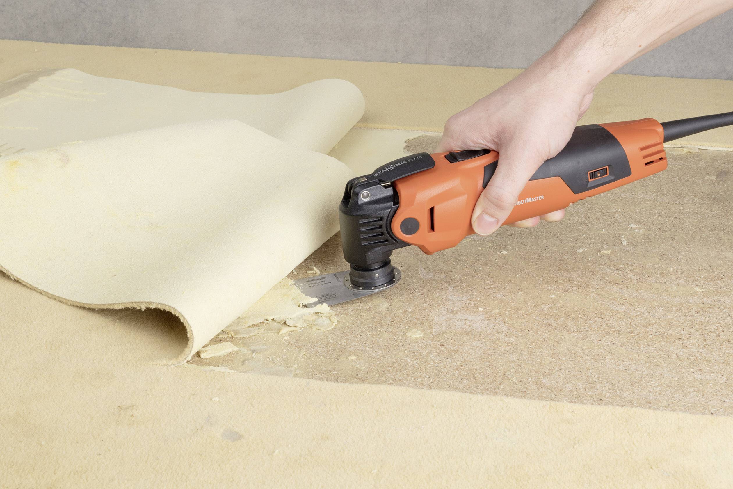 A person is using an orange electric tool to remove carpet from a wooden floor.