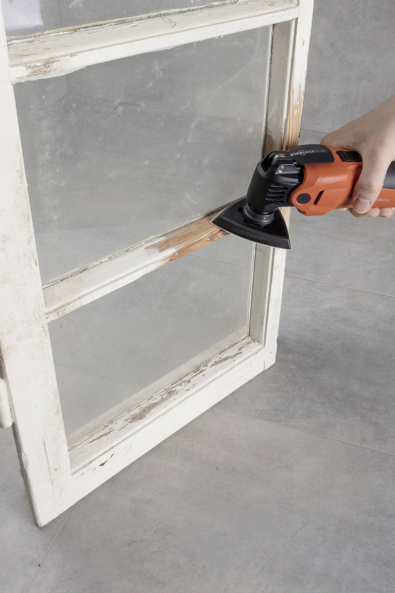 A person is sanding old paint off a wooden window frame using an electric sanding tool. The frame is being renovated.