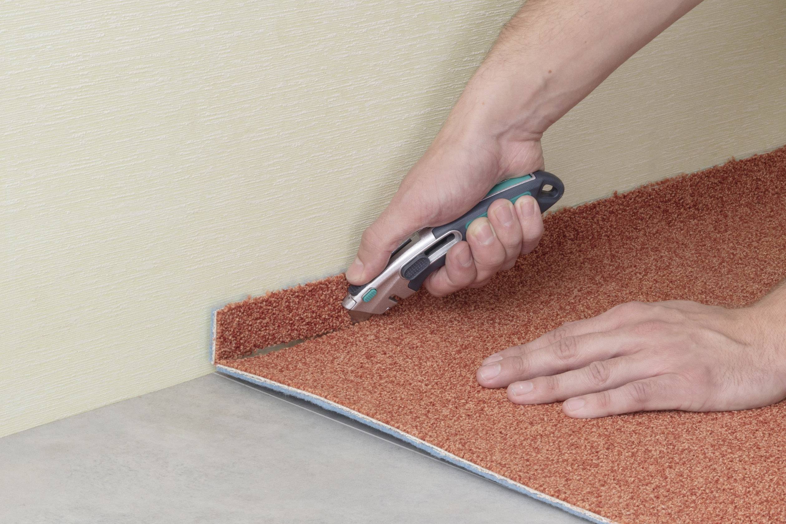 A person is cutting a red-brown carpet along a wall using a craft knife. Hands are visible.