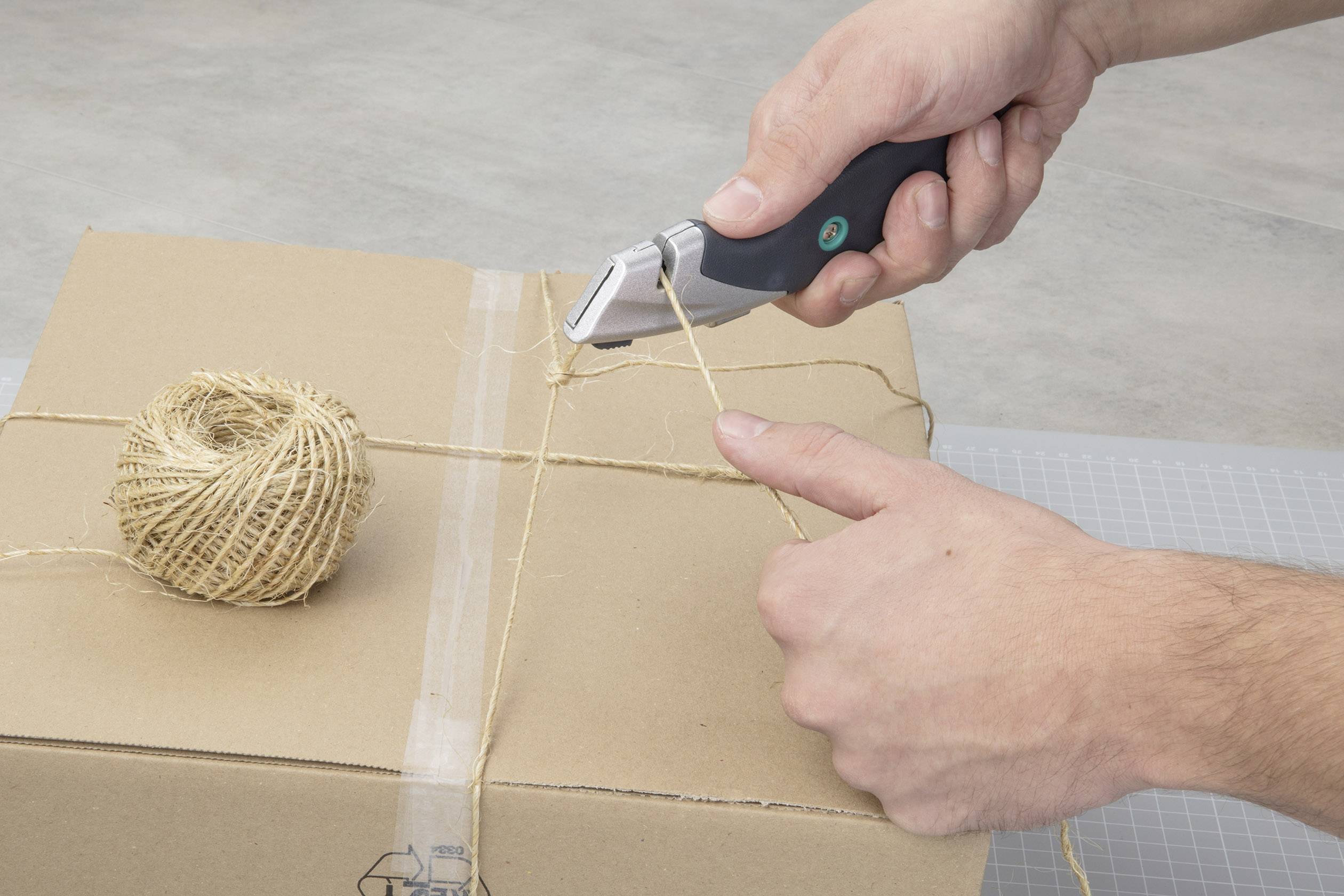 Two hands are cutting a piece of string with a knife on a cardboard box, with a reel of coarse twine lying beside it.