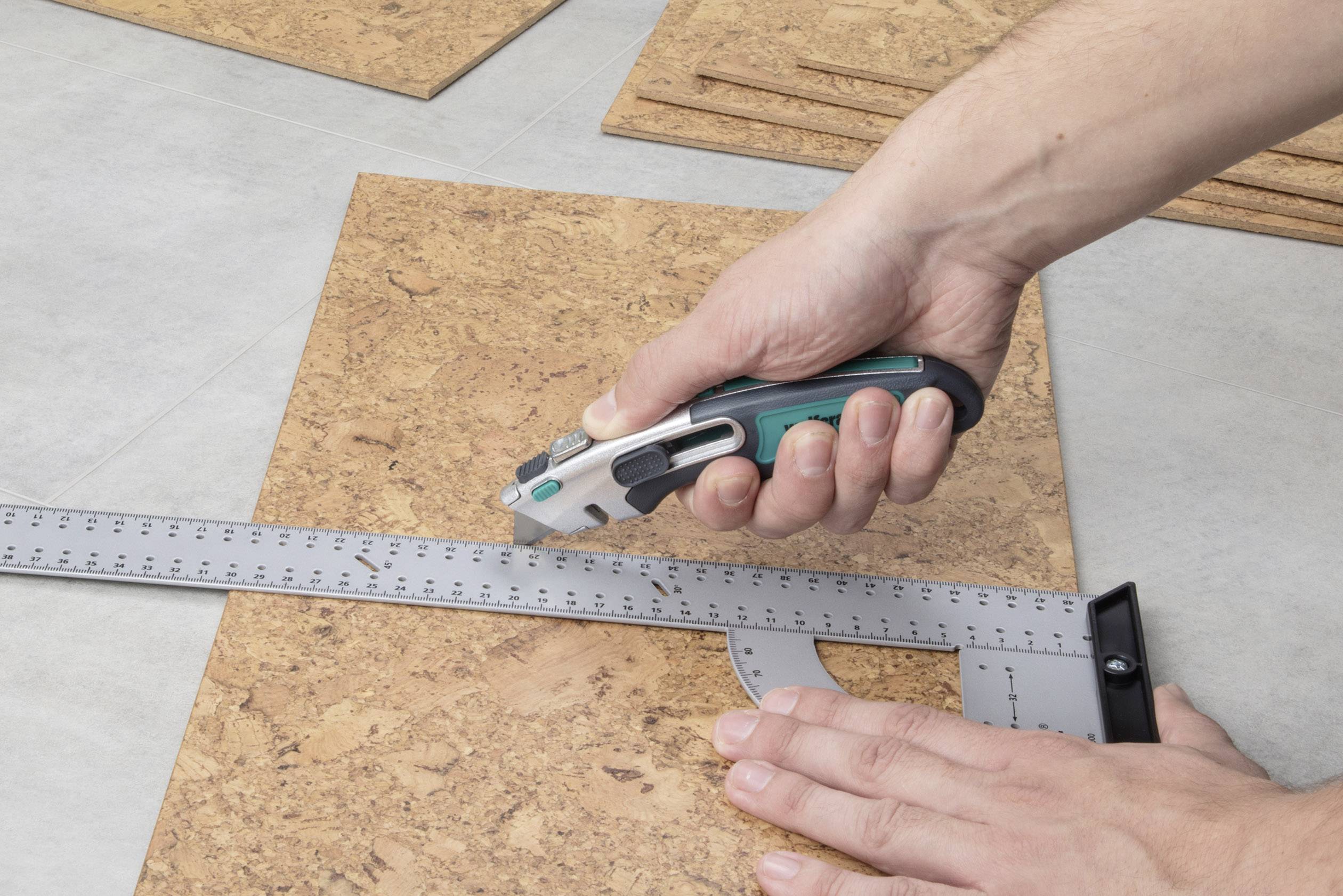 A person is cutting along a metal angle using a craft knife on a cork board. Hands are holding the knife and angle.