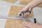 A person is cutting along a metal angle using a craft knife on a cork board. Hands are holding the knife and angle.