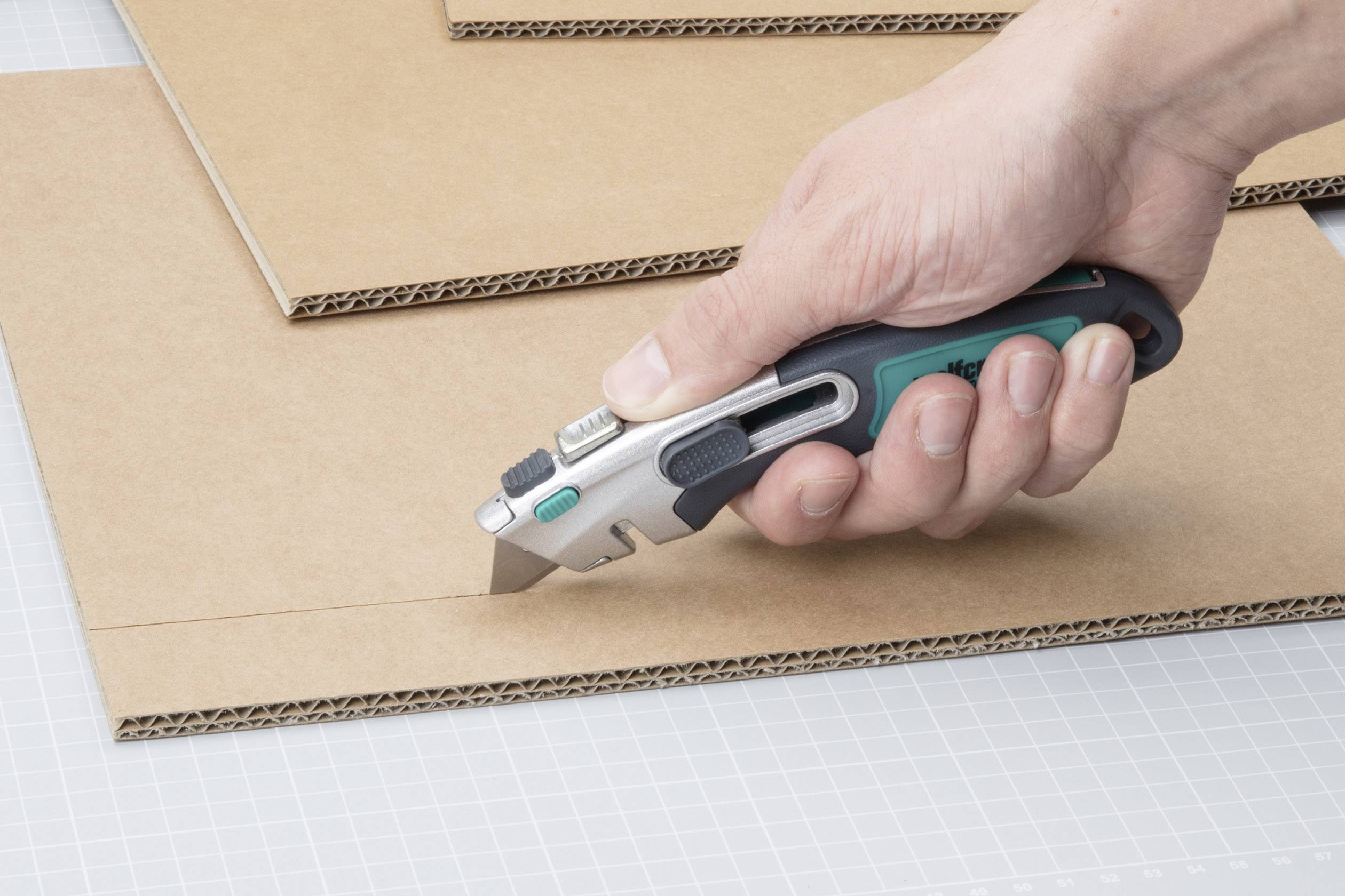 A hand cuts through cardboard with a craft knife on a chequered surface.
