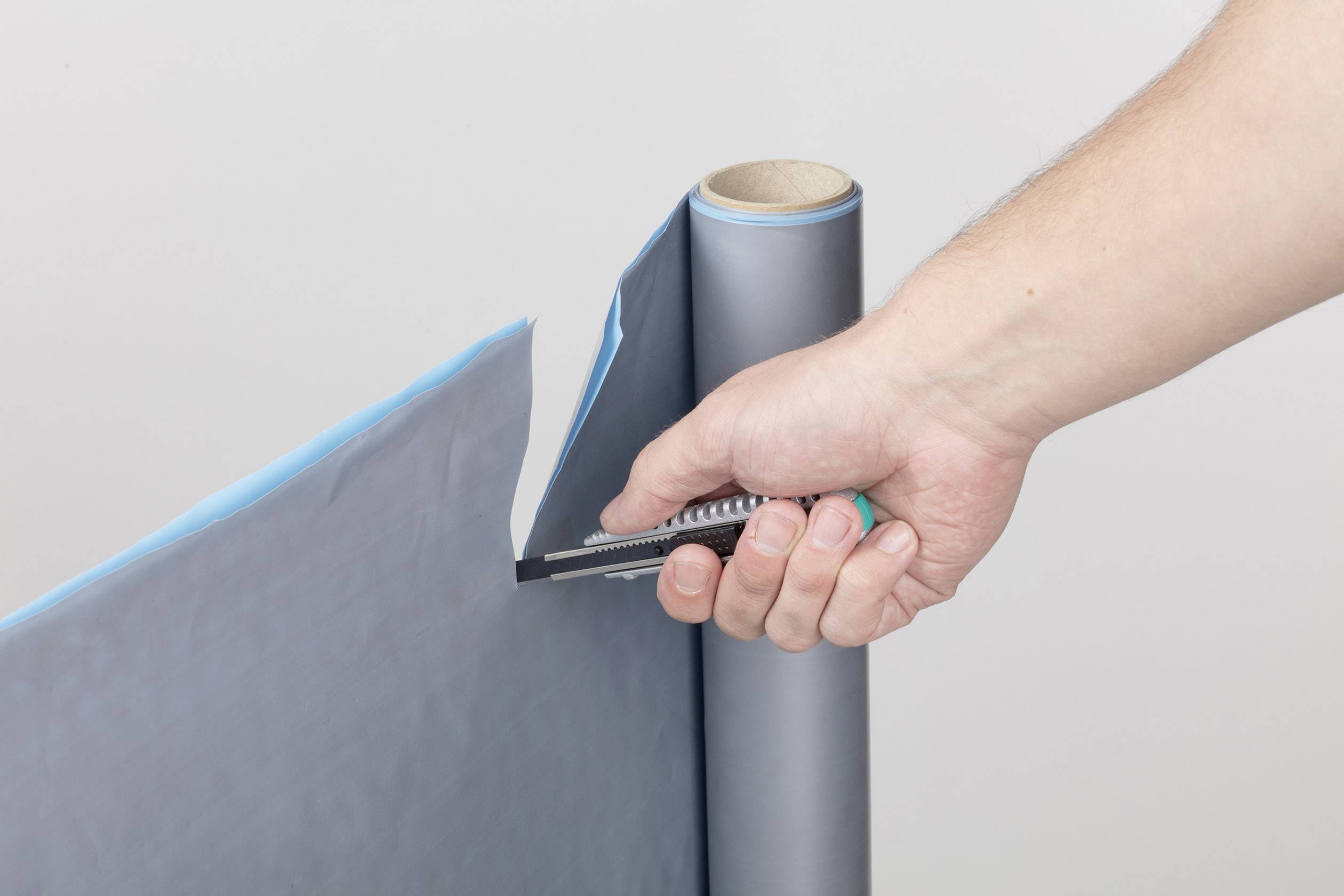 'Person cuts grey film from roll using a stanley knife.'