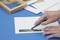 A hand is cutting a rectangular section with a craft knife into white paper on a blue cutting mat, with a ruler and frame nearby.