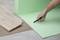 A hand cuts a green underlay for laminate flooring with a utility knife. Laminate planks are laid out nearby.