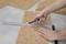 A person is cutting a cork board with a utility knife, supported by a metal ruler, on a grey floor covering.