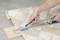 A person is cutting a beige tile floor covering with a craft knife and a metal ruler on a wooden board.