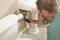 A man is examining a pipe beneath a sink with a measuring device. Next to the sink is a toilet.