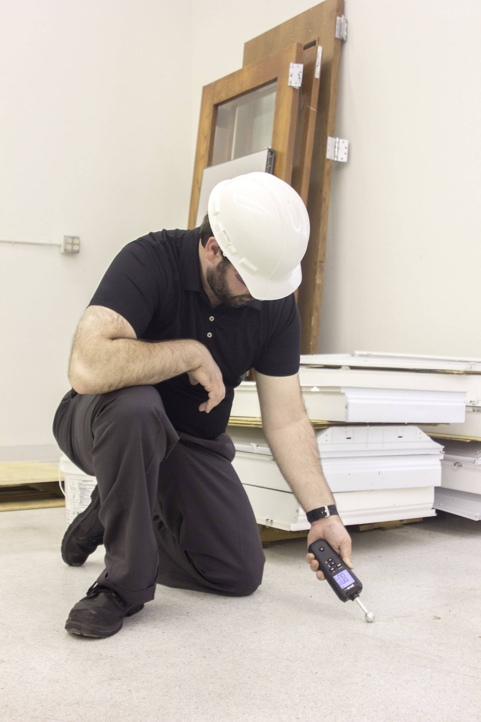 A man wearing a white hard hat is measuring the moisture content of the floor in a room where stacked door frames are visible.