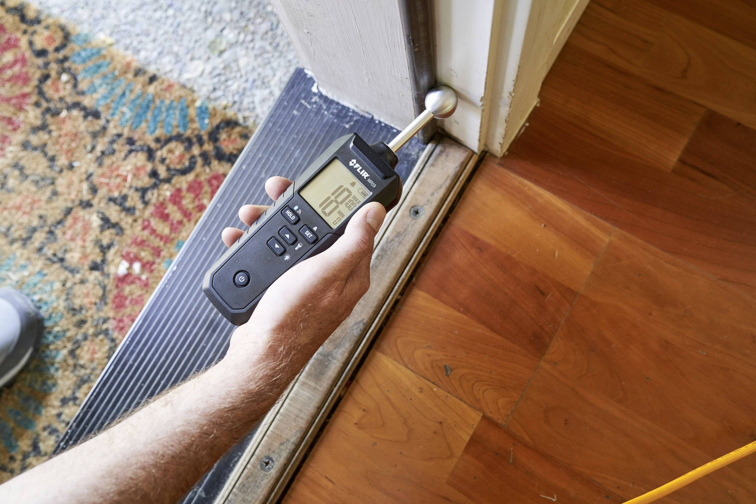 A person is measuring soil moisture near a doorstep using a digital device. The value is displayed on the screen.