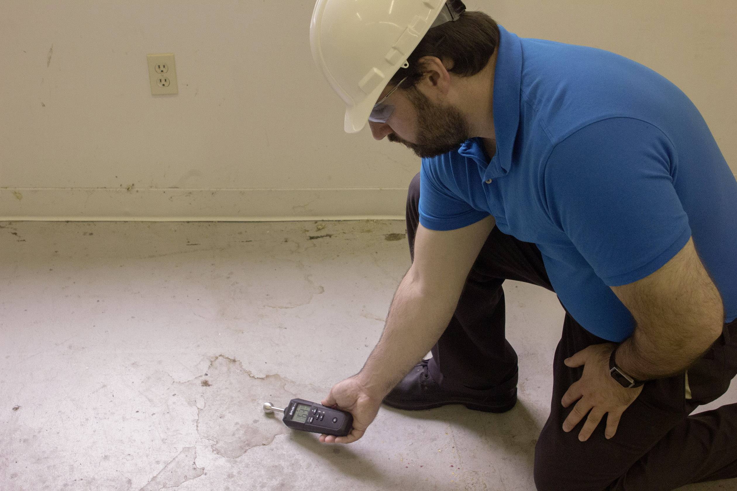A man wearing a hard hat and safety glasses is measuring moisture on a concrete floor using a portable device.