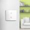 A modern, white light switch with two red touch buttons on a grey wall. A sofa and shelving unit are blurred in the background.