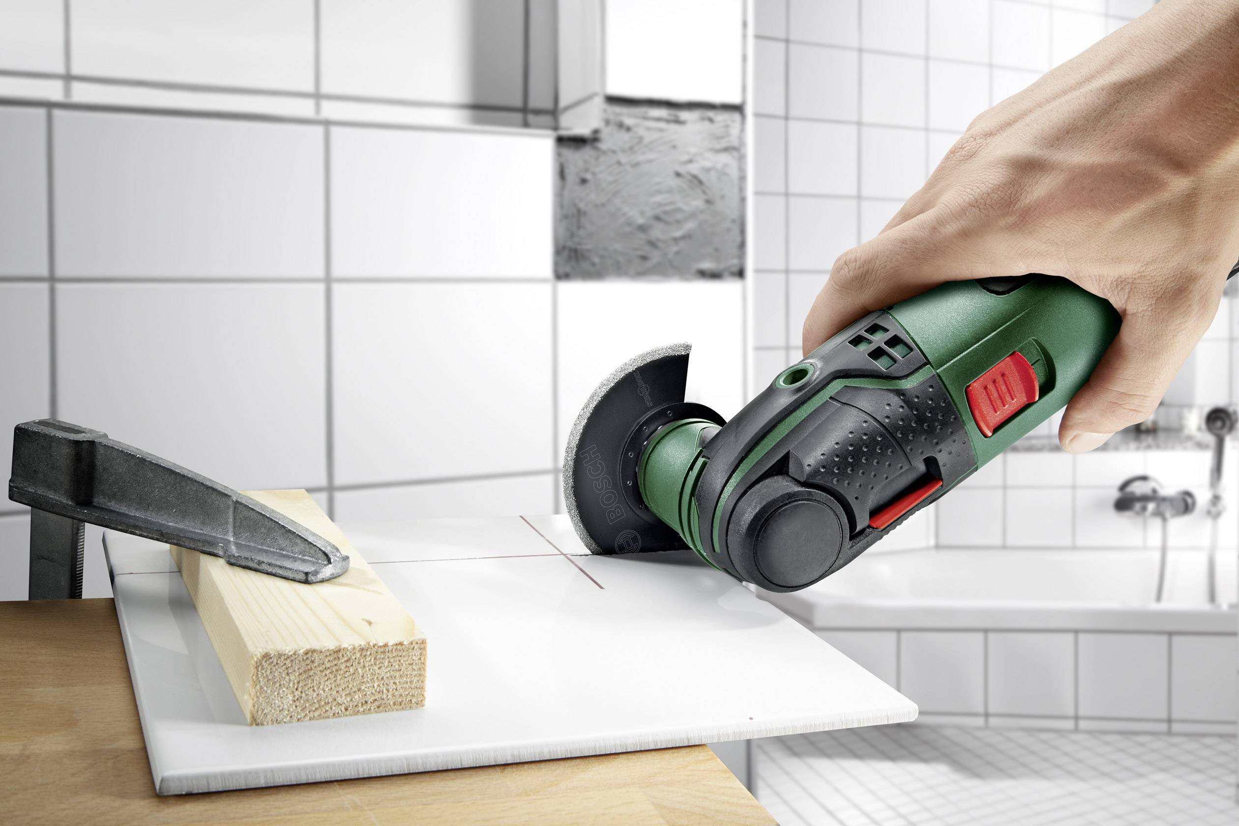 A hand is holding a green electric tool cutting a tile on a wooden block. A tiled bathroom is visible in the background.
