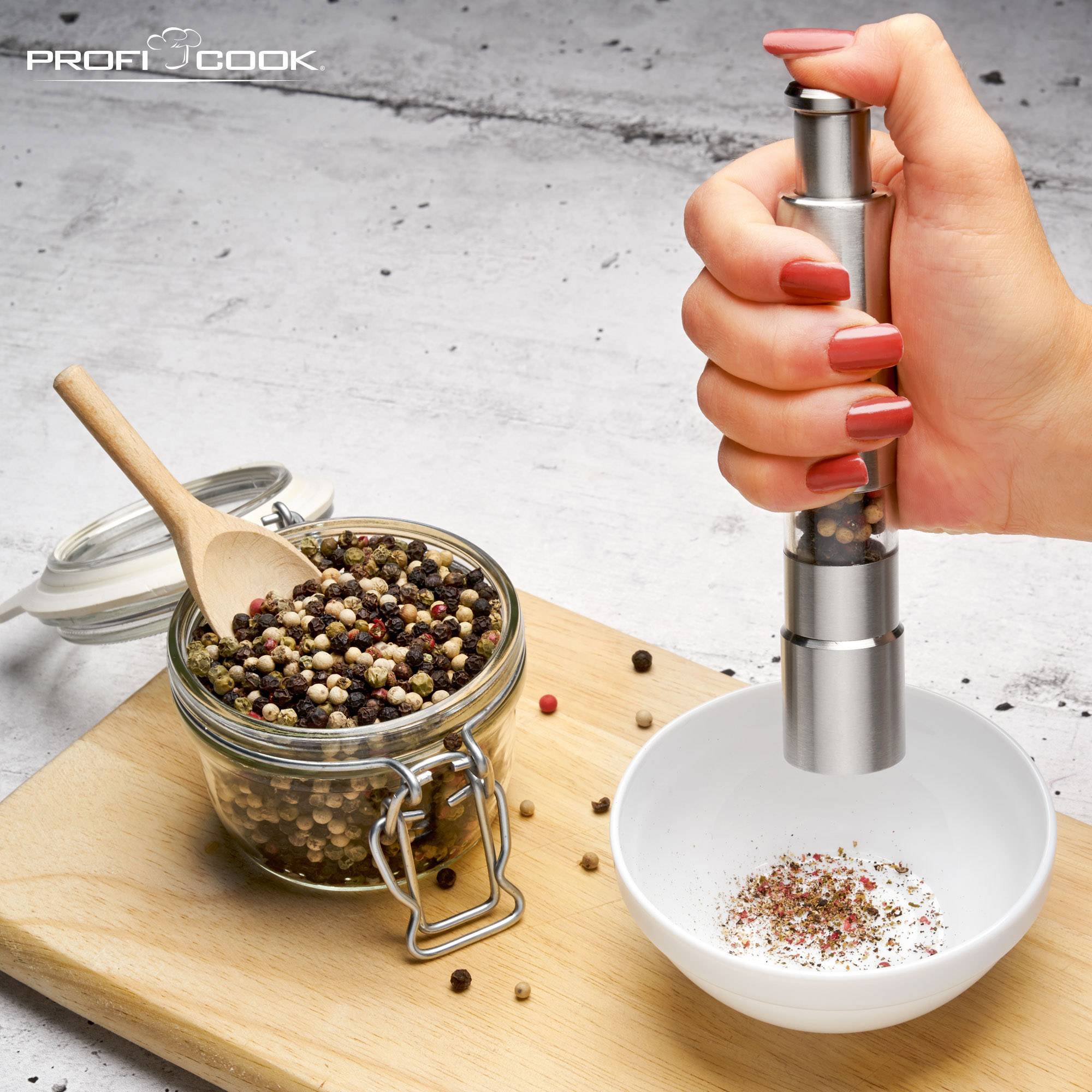 A hand is holding a pepper mill over a white bowl. To the right, there is a glass with colourful peppercorns and a wooden spoon.