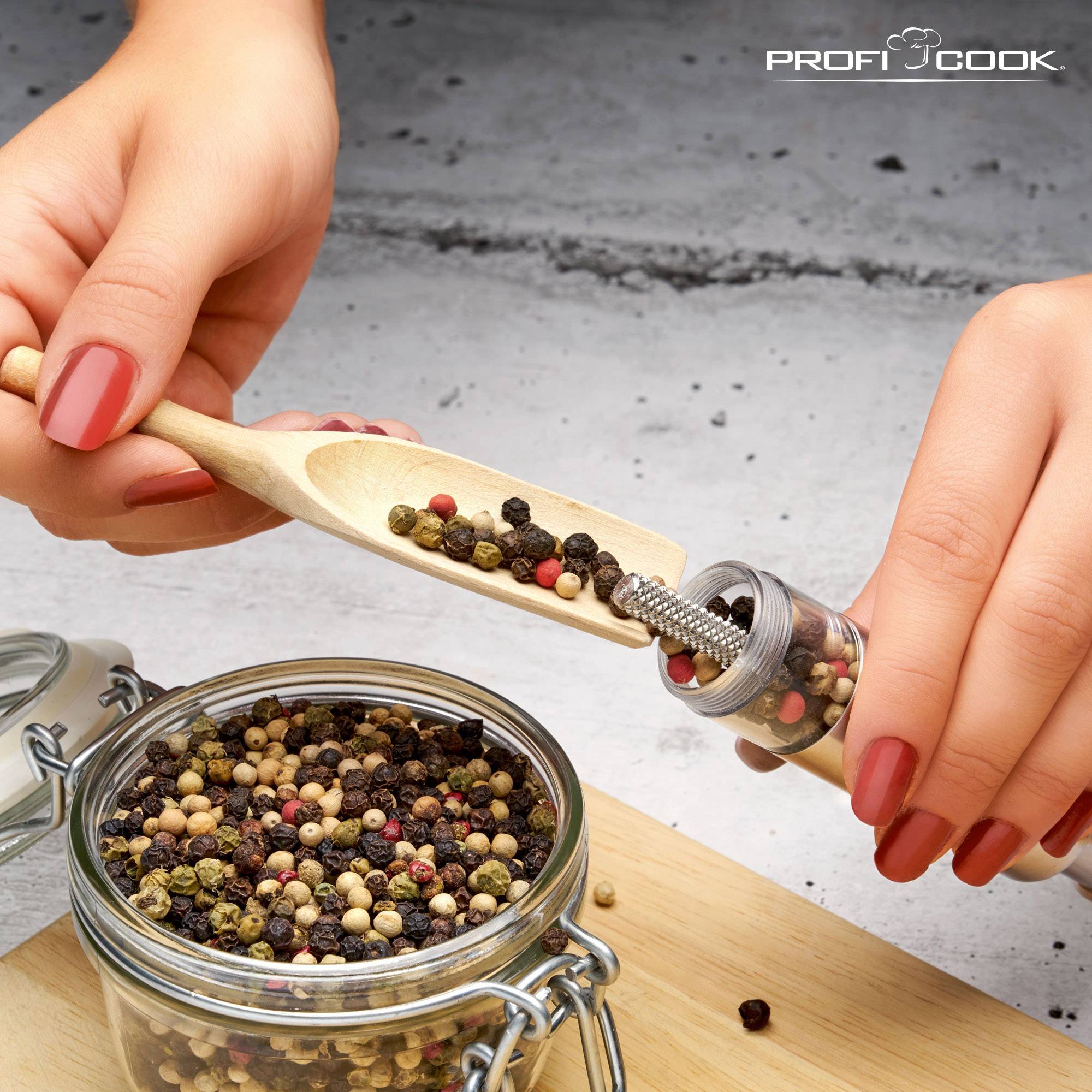 A hand fills a transparent pepper mill with colourful peppercorns using a wooden spoon, positioned over an open storage jar.