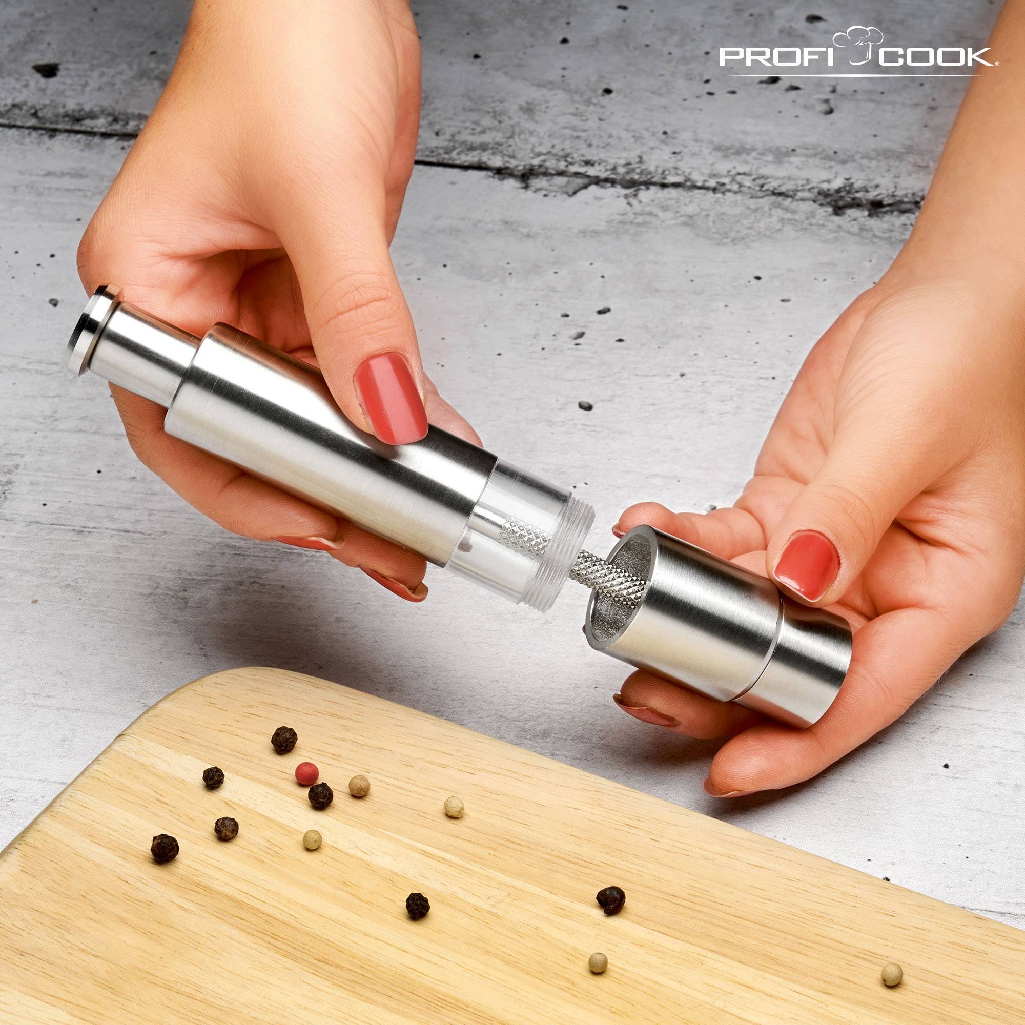 A stainless steel pepper mill is opened to grind peppercorns. Various peppercorns are lying on a wooden board.