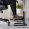 A leg in a brown shoe resting on an ergonomic footrest under an office chair next to a small table with plants.