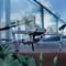 A grey drone with a camera flies just above a plant surface against a blurred city or building background.