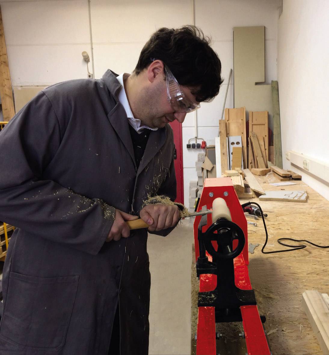 A person is using a wood grinding machine in a workshop, wearing safety glasses and an overcoat. Wood shavings are falling to the floor.