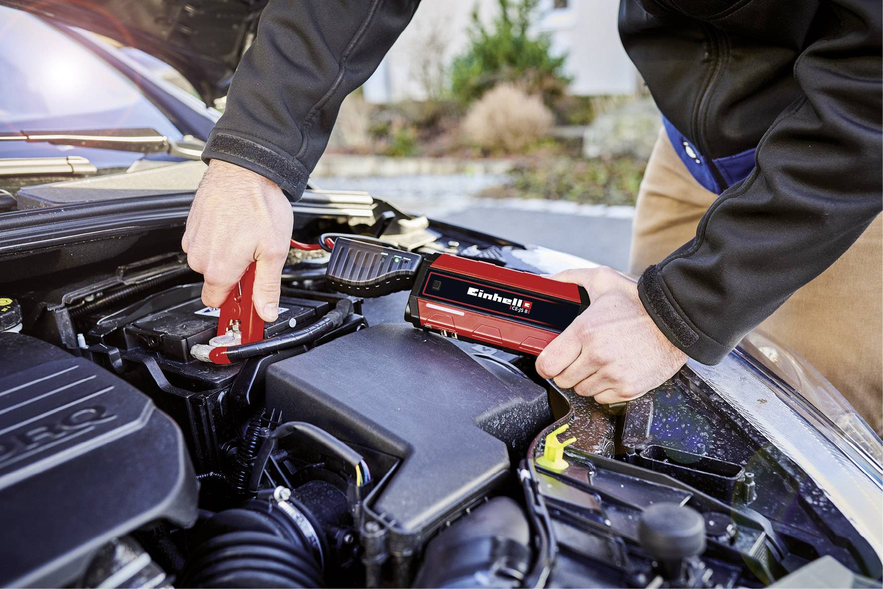 A person is using a portable jump starter to jump-start a car battery. Red and black clamps are connected.