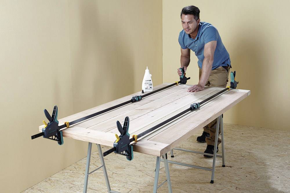 A man is working with wood on a workbench using clamps and wood glue in a room with a light wooden floor and beige walls.