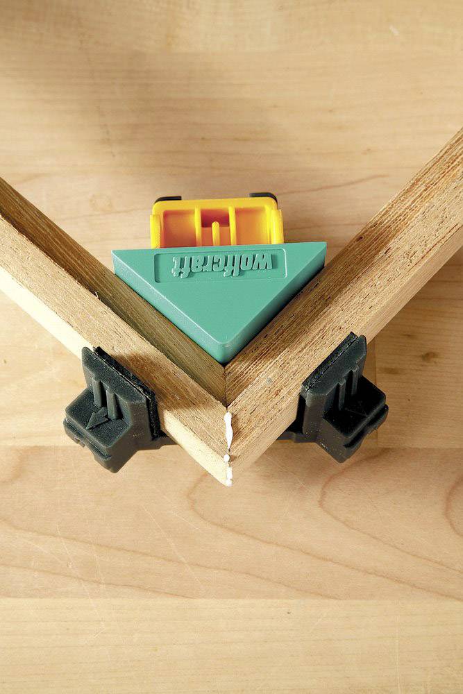 A green angle clamp holds two pieces of wood together at a 90-degree angle on a table.