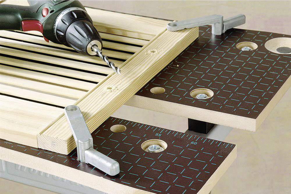 A drill is lying on a workbench and drilling holes into a wooden board secured with clamps.