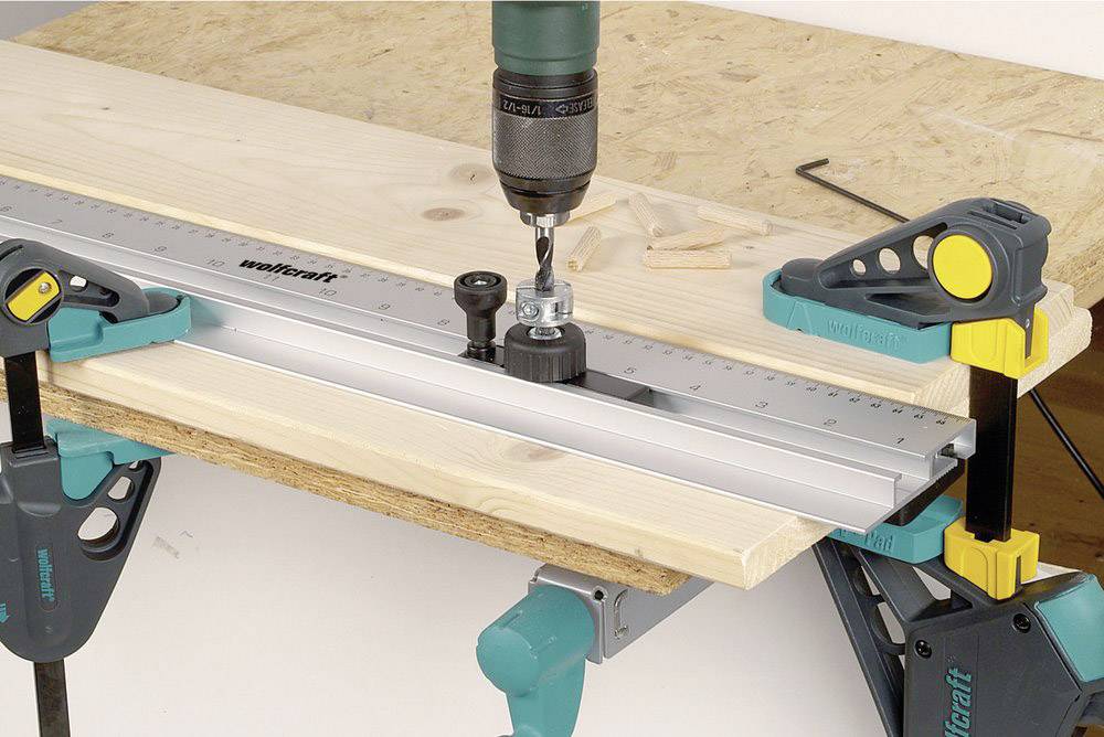 A piece of wood is secured to a table using clamps. A drill is attached to a ruler to perform precise drilling.