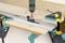 A piece of wood is secured to a table using clamps. A drill is attached to a ruler to perform precise drilling.