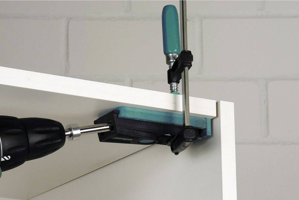 A drill creates holes in a white shelf board secured to a workbench using a G-clamp.