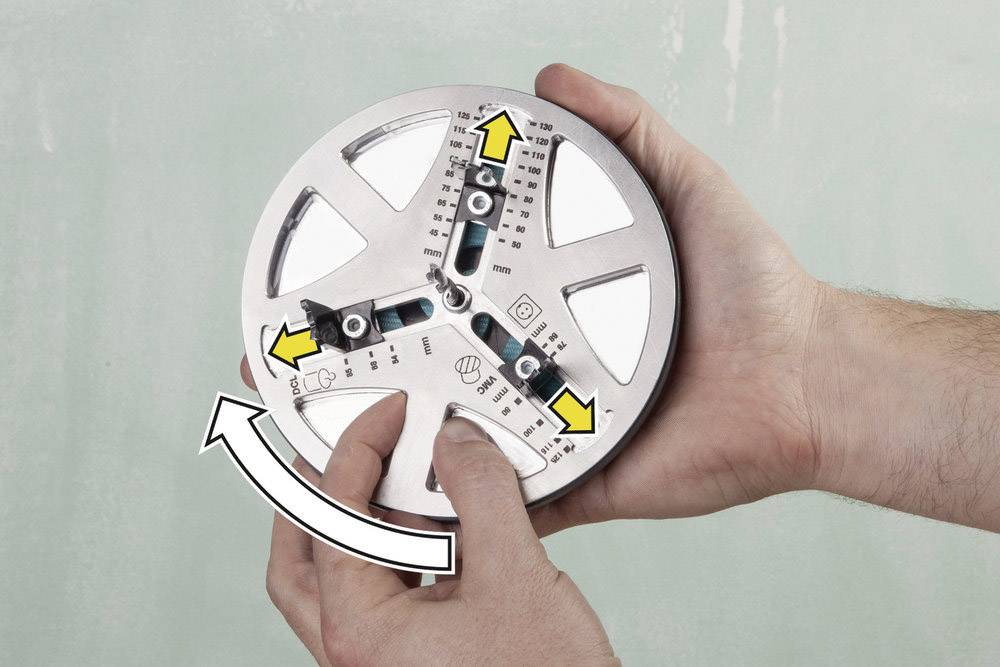 Two hands are holding a round metal tool, whose arrows and markings indicate settings and adjustments.