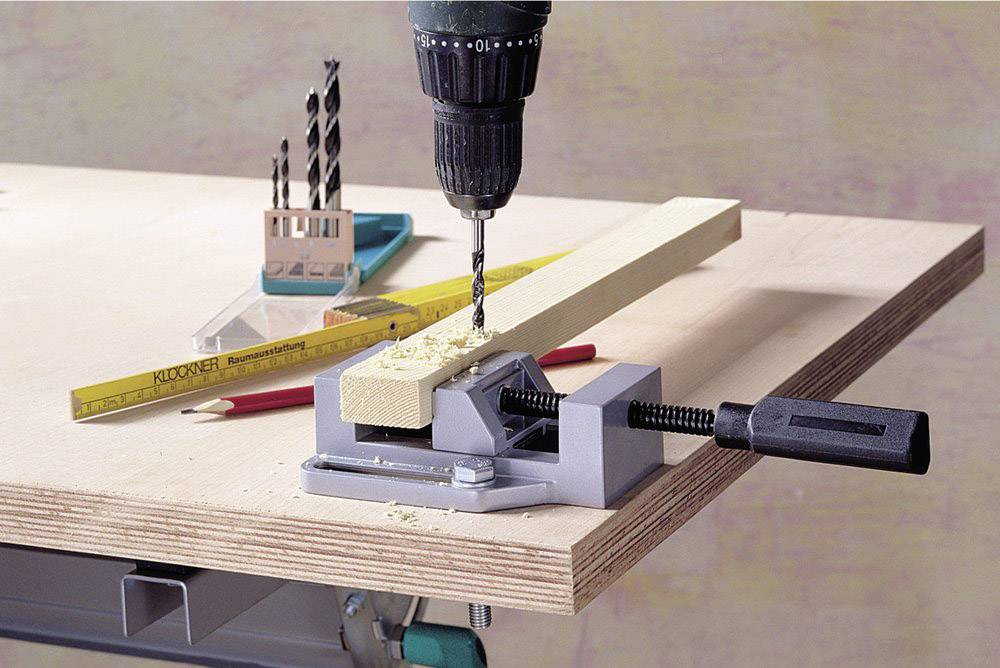 An electric drill is boring a hole into a piece of wood, which is secured in a vice on a table. Drill bits are lying nearby.