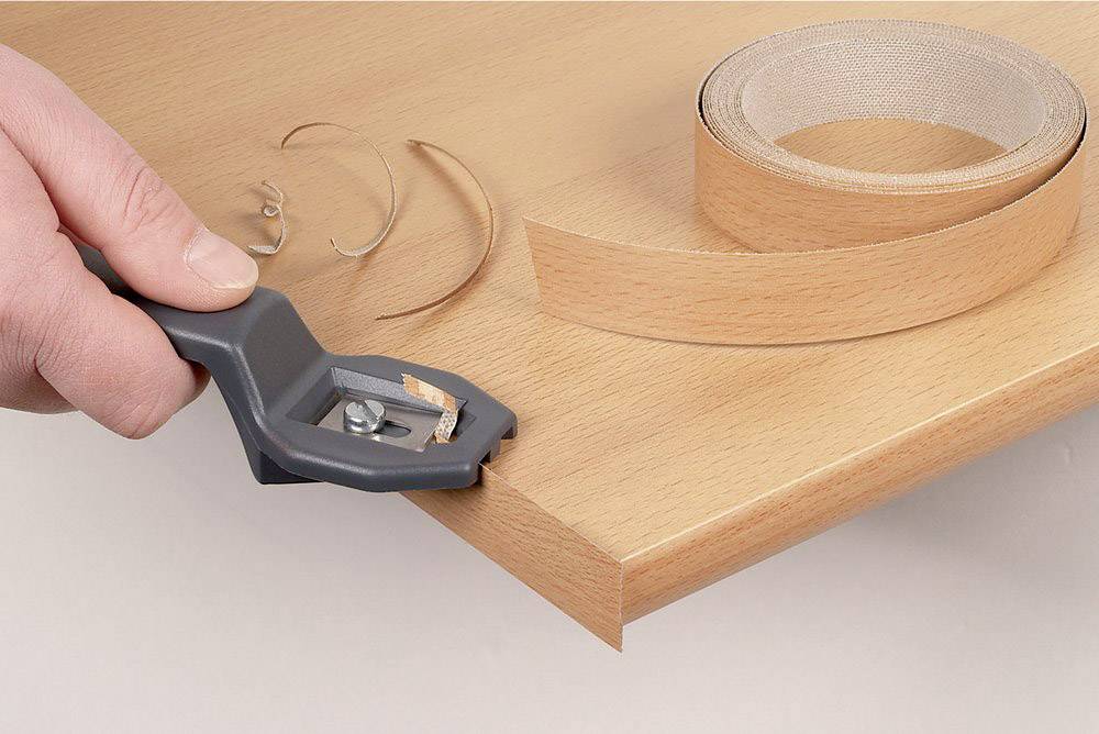 A hand is using an edge banding machine to apply a veneer edge to a wooden board. A roll of veneer lies beside it.