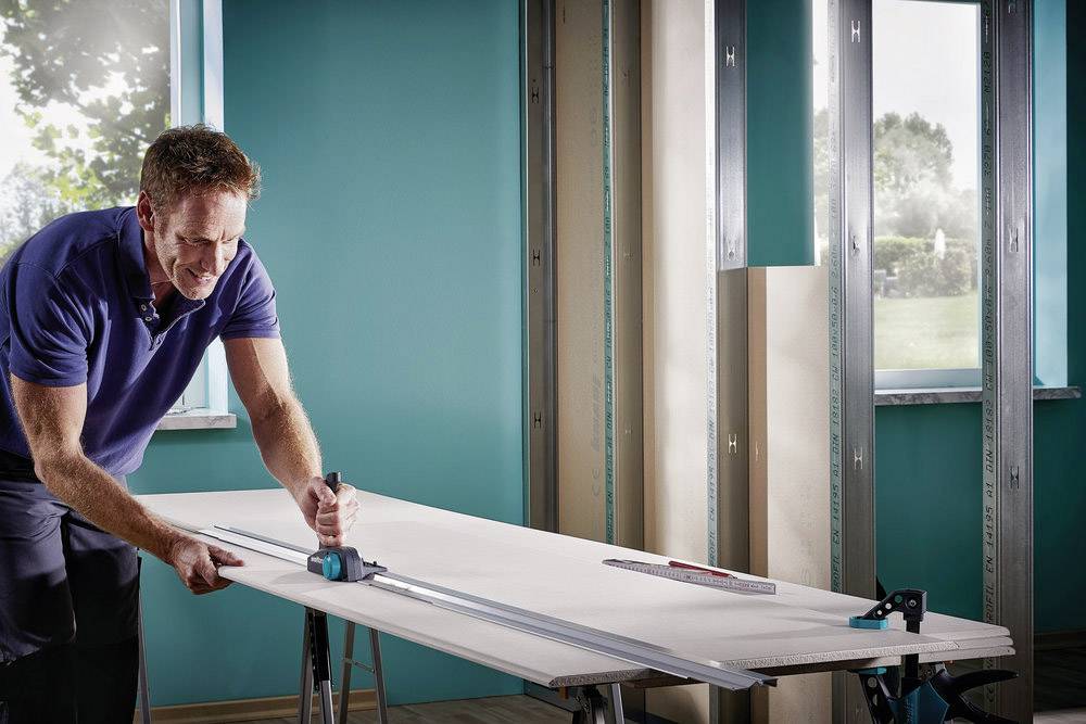 A man is cutting a plasterboard with a mitre saw in a room with turquoise walls, daylight streaming through the window.