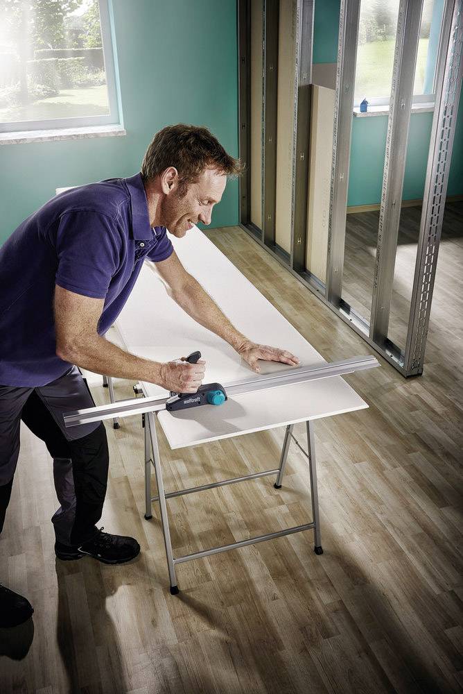 A man is measuring and cutting a white board in a room with a wooden floor and unfinished walls, with light streaming through a window.