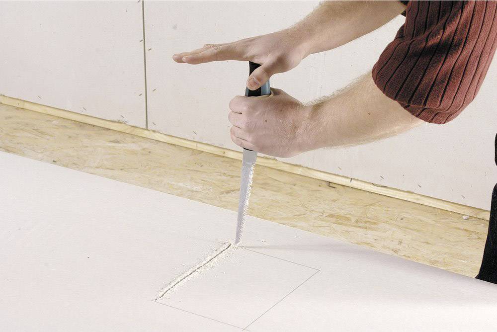 A person is cutting a rectangular hole in a white plasterboard using a saw.