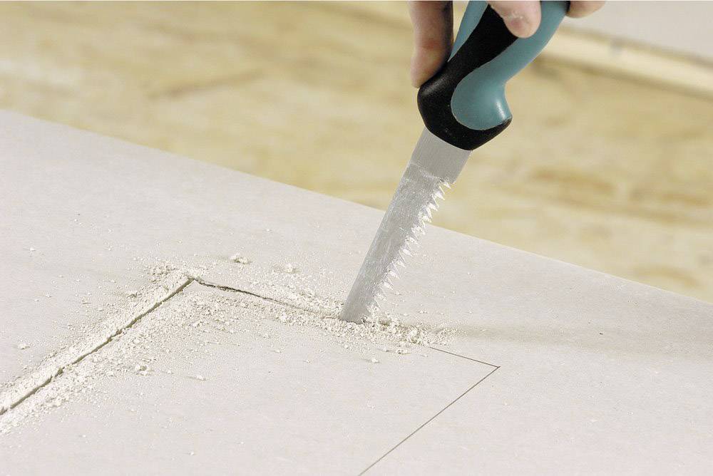 A hand holds a saw and cuts a rectangular hole in a plasterboard. Sawdust is scattered around the cut.