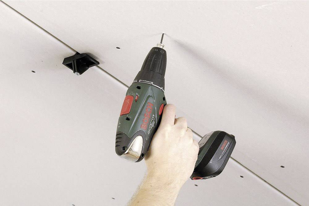 A hand is holding a cordless drill and screwing a screw into a ceiling panel. The drill is black and green.
