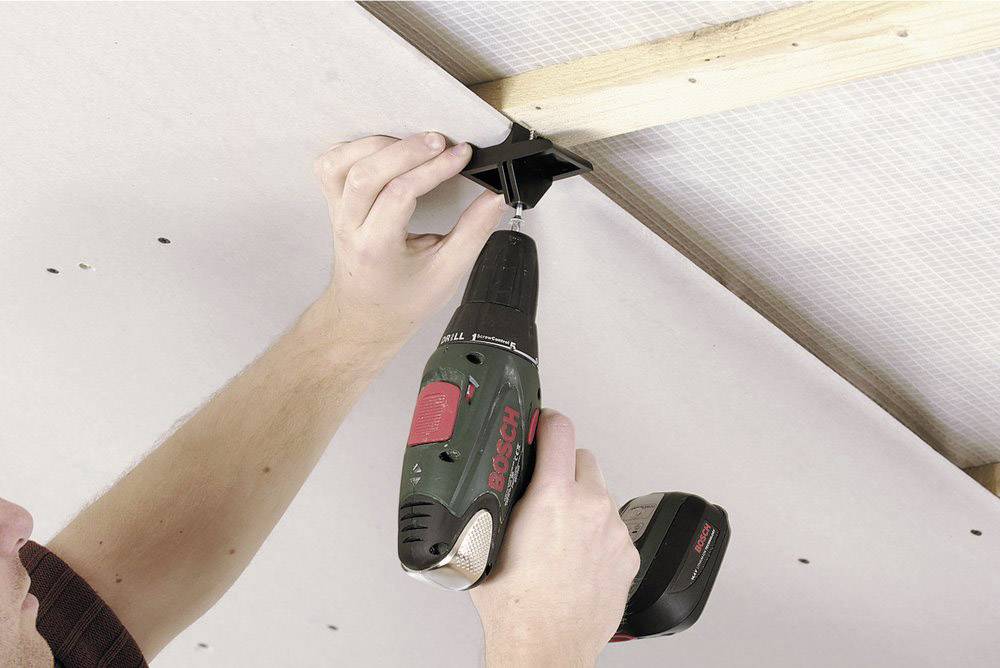 A person is securing a black mounting bracket to a light-coloured ceiling using an electric screwdriver.