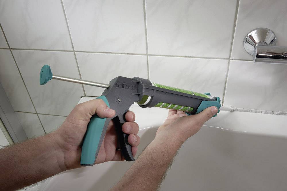 A person is using a cartridge gun to apply sealant to the joints between a bath and white tiles.