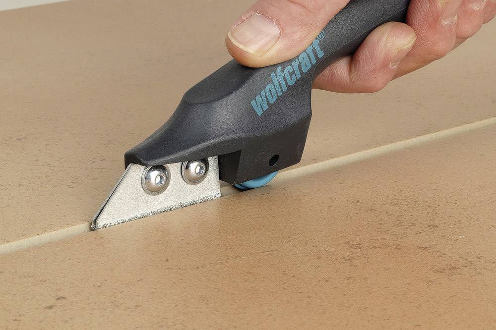 A hand is holding a tile cutter, scoring a groove in a tile to split it.