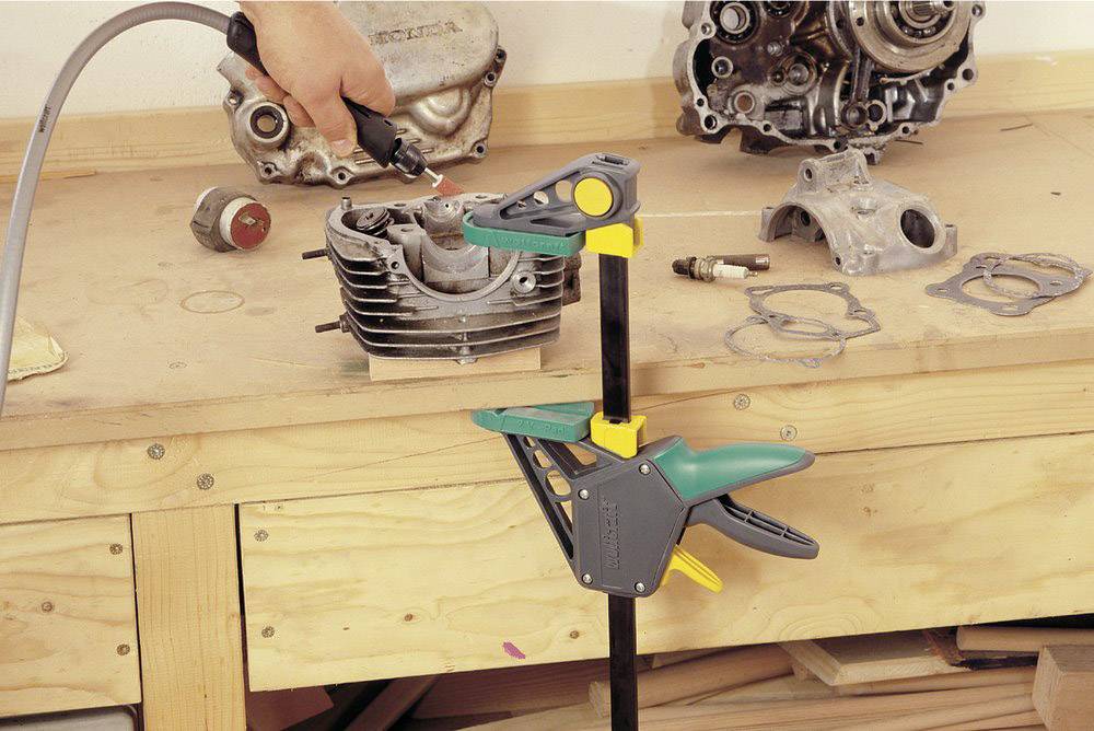 A hand is using a pneumatic tool to machine a motor piston on a workbench. Various motor parts are lying beside it.