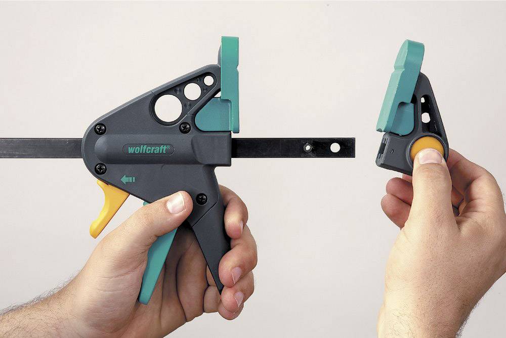 A hand is holding a one-handed plastic clamp, the second hand holding a removable trigger release. Clamp in grey and green design.