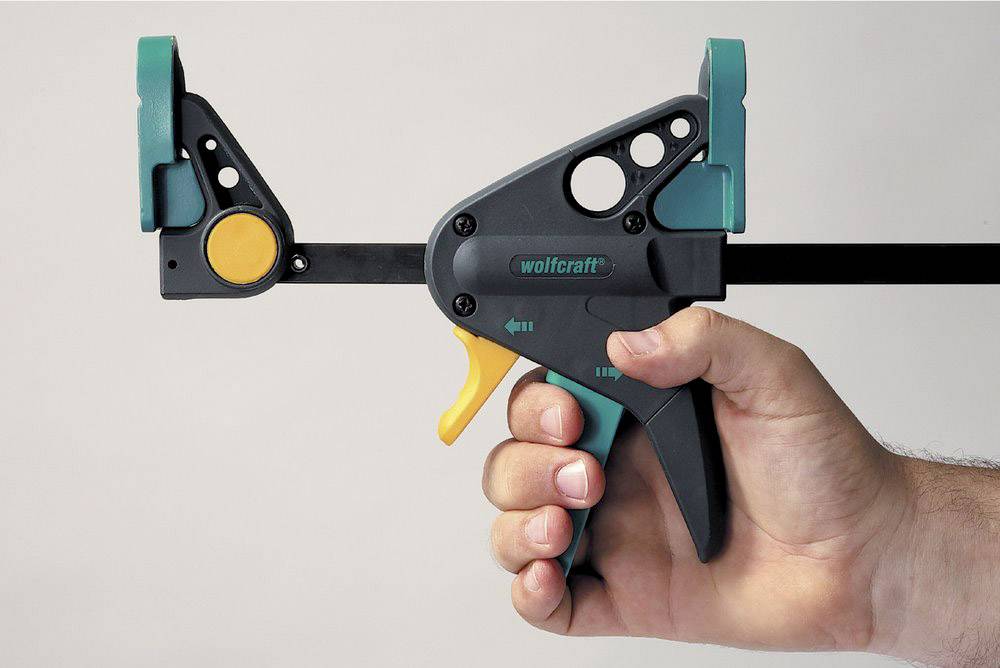 A hand is holding a green and black one-handed G-clamp with a yellow lever from Wolfcraft. The clamp is open and ready for use.