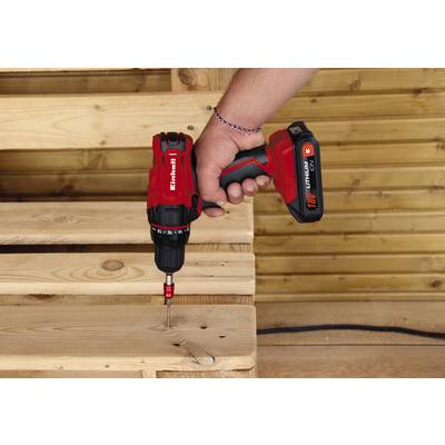 A hand is holding a red cordless drill and screwing a screw into a wooden pallet. The background is a wooden panelling.