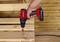 A hand is holding a red cordless drill and screwing a screw into a wooden pallet. The background is a wooden panelling.