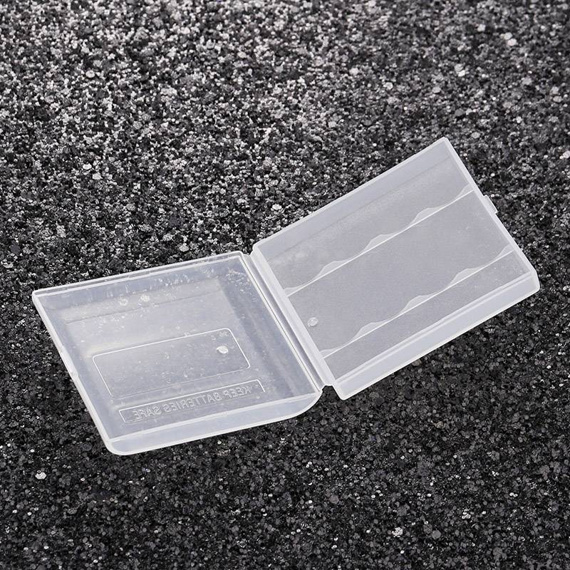 Transparent, empty plastic battery holder on a grey-black textured surface. The holder is open, revealing battery compartments.