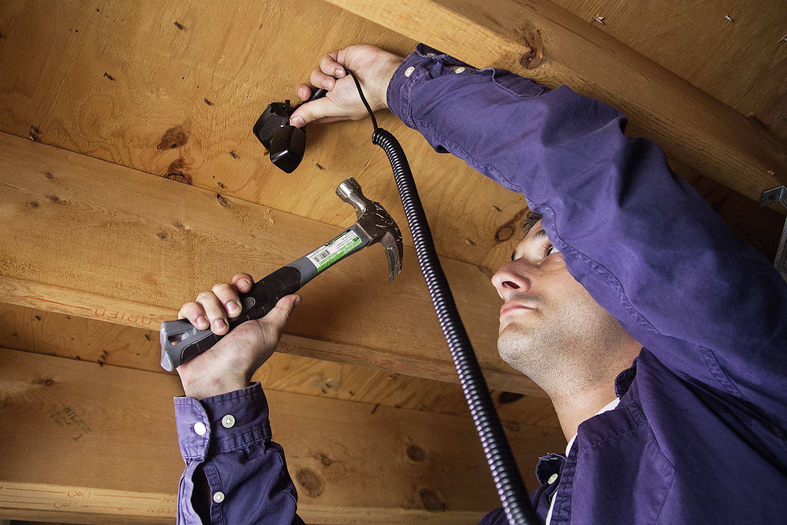 A man in blue clothing is fixing a camera to the wooden ceiling of a building using a hammer. He is holding the camera with his other hand.
