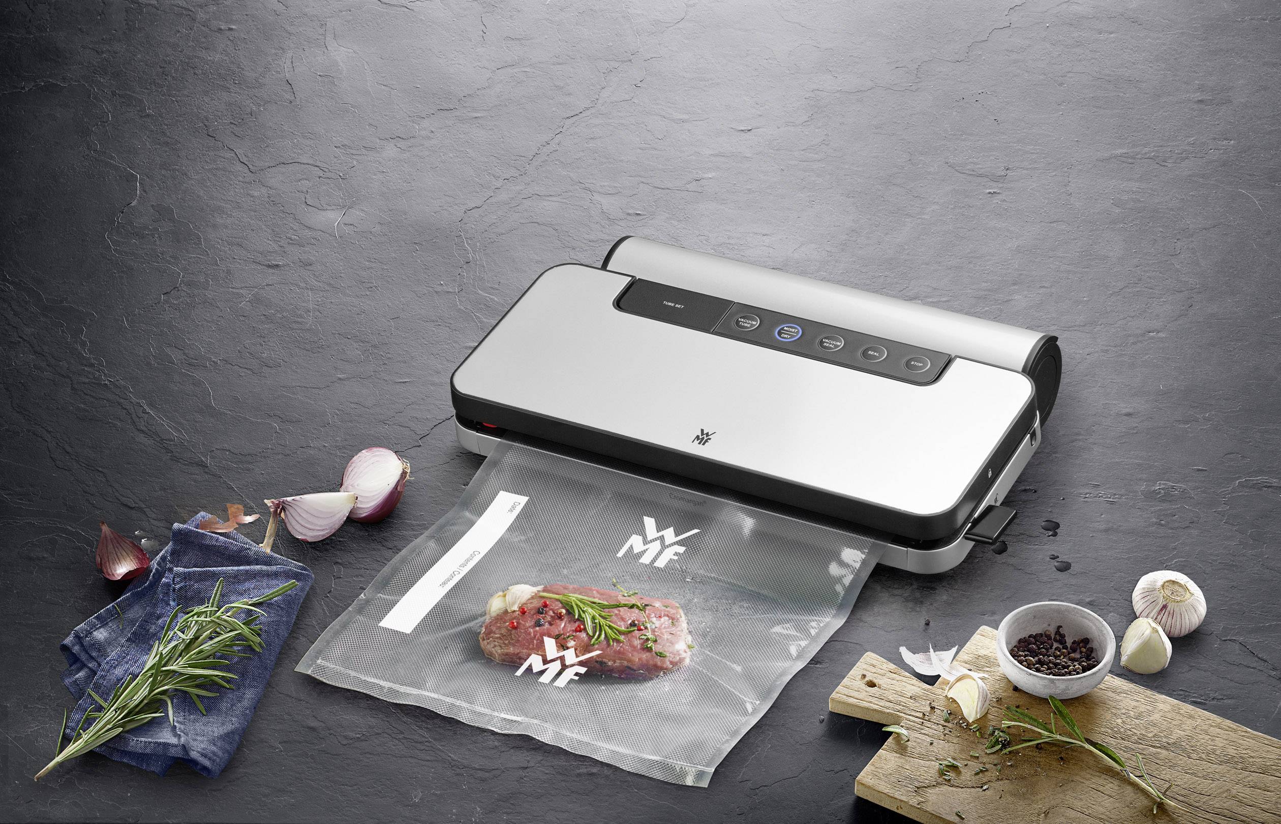 WMF vacuum sealer seals a piece of meat with spices in a plastic bag. Onions, garlic, and rosemary are lying on the table.